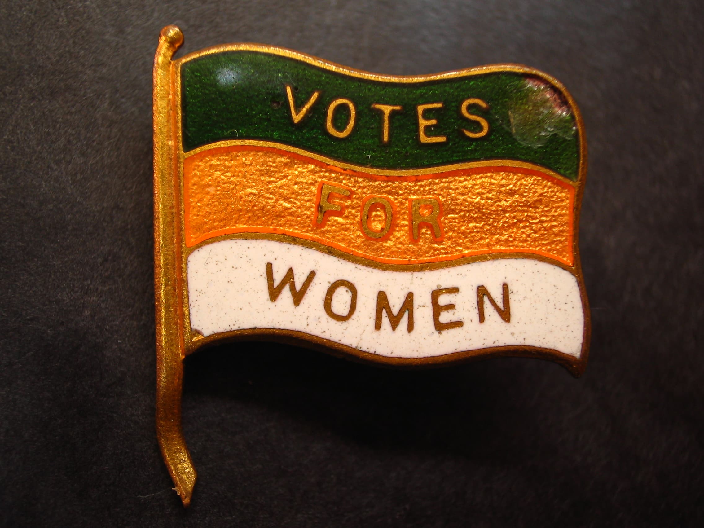 A pin with the colours of suffrage saying votes for women