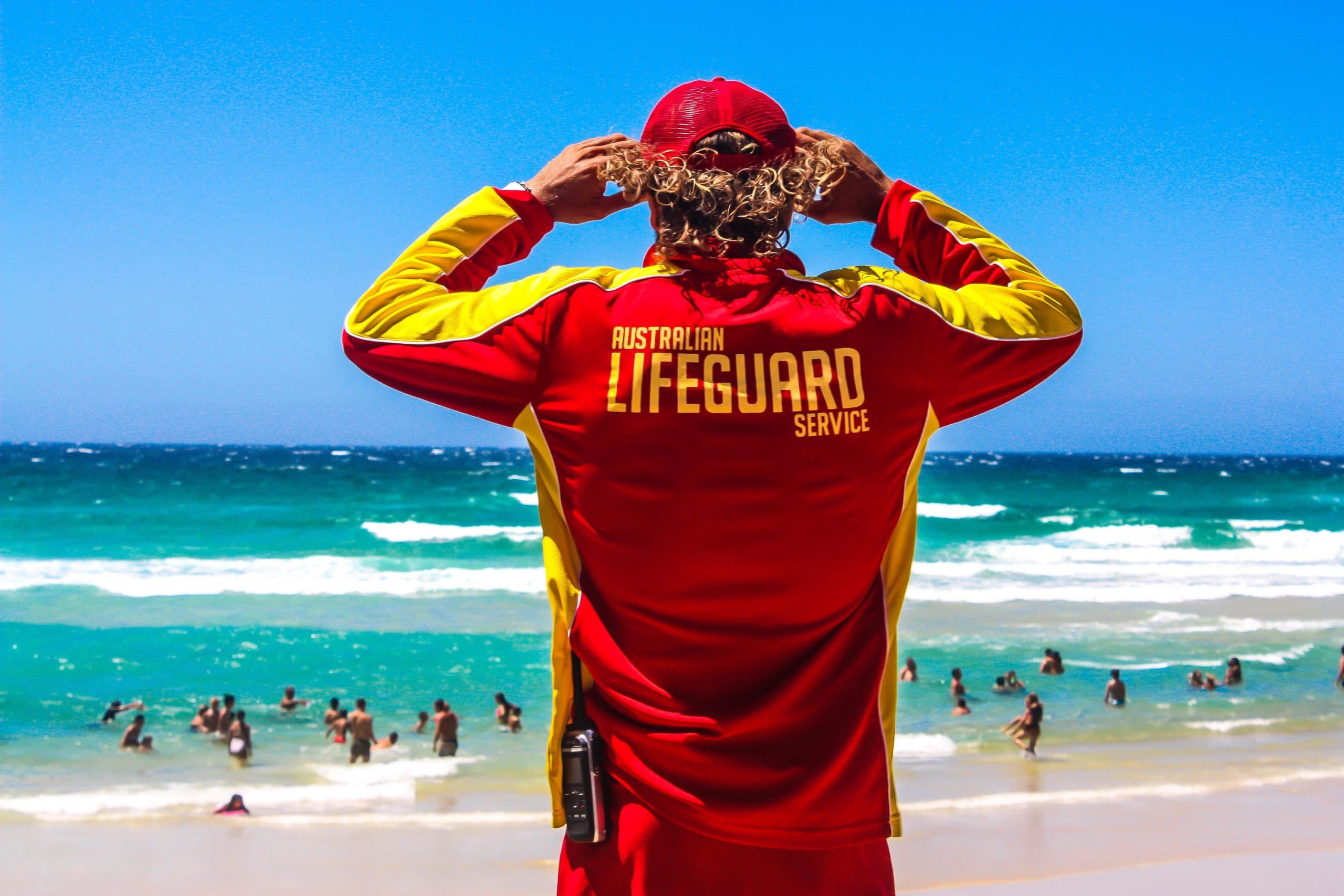 Lifesaver wearing red and yellow uniform standing on the beach, looking out to sea.