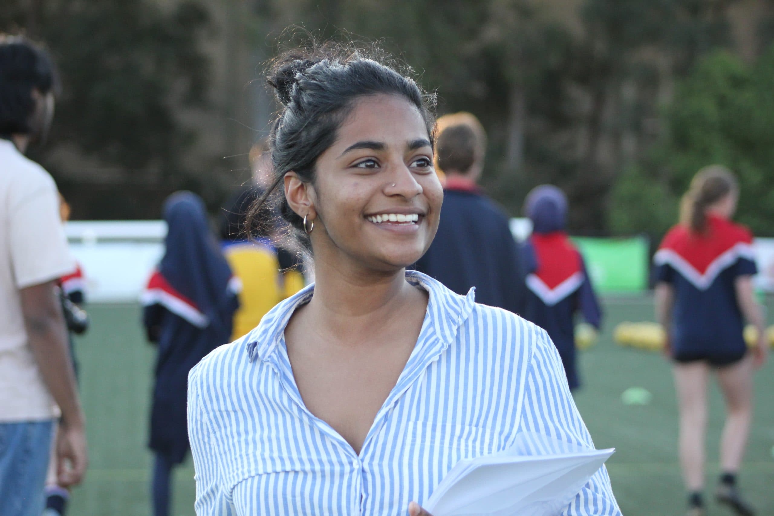 A young woman smiling and holding a pile of papers in one arm. She is standing outdoors at a friendly football match.