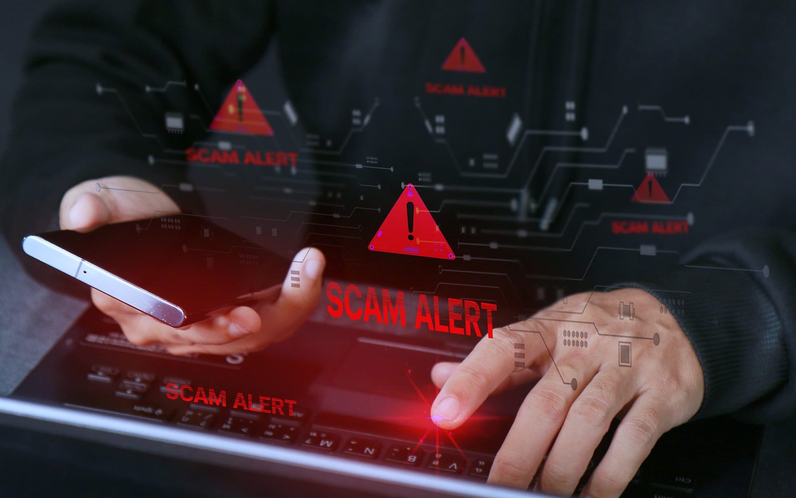 A dimly lit man's hands poised over a keyboard and holding a phone in one hand. A scam alert warning sign is superimposed.