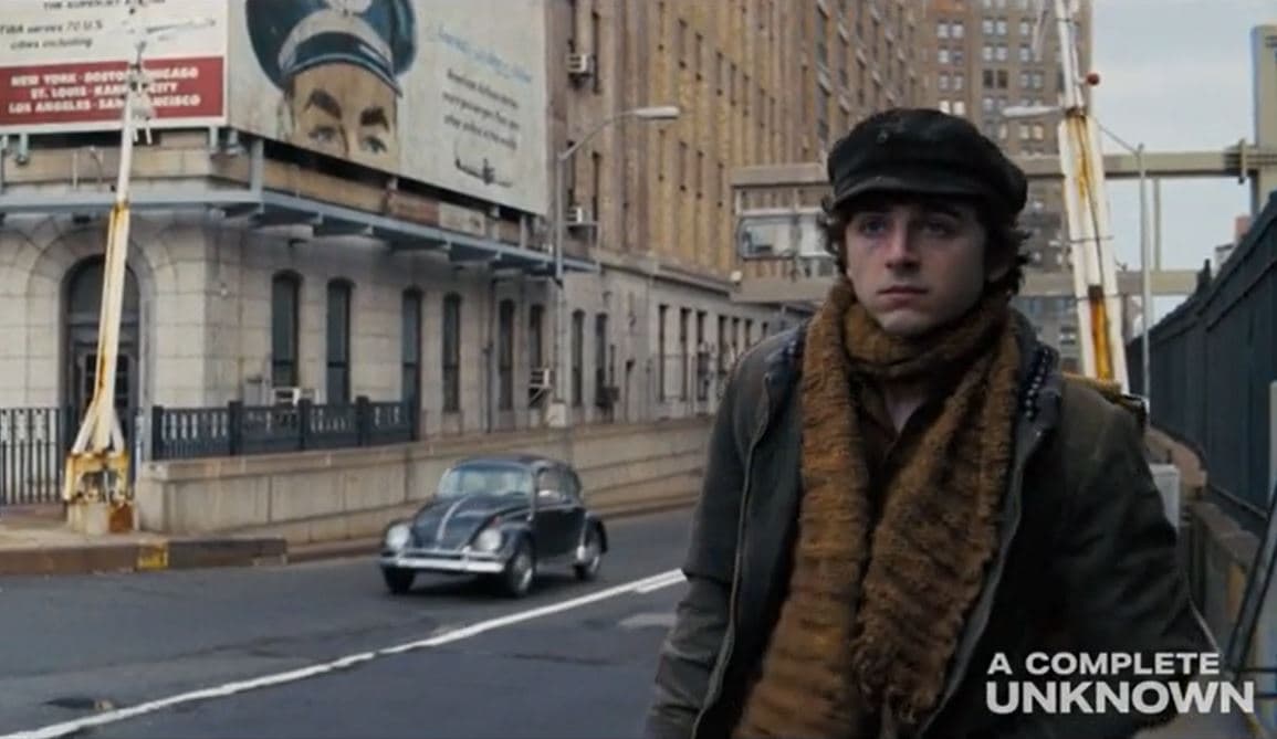 Frame from the movie A Complete Unknown showing a young man carrying a guitar down a busy city road.
