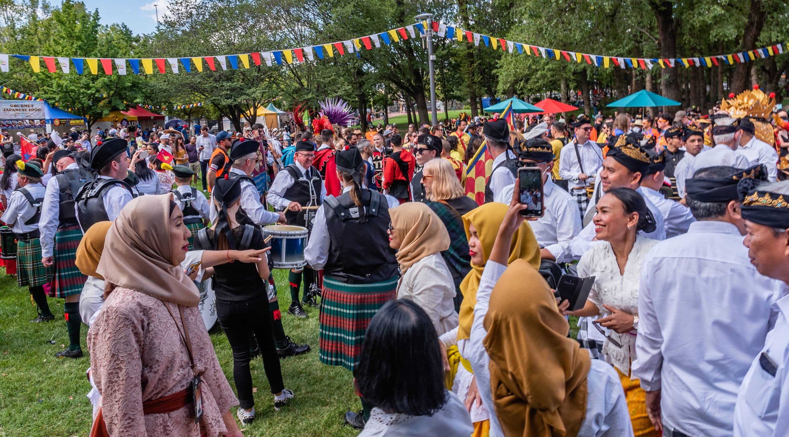 National Multicultural Festival in Canberra 2020. Large group of people in many types of cultural dress, some playing music. They are outdoors in a park with stalls and bunting.