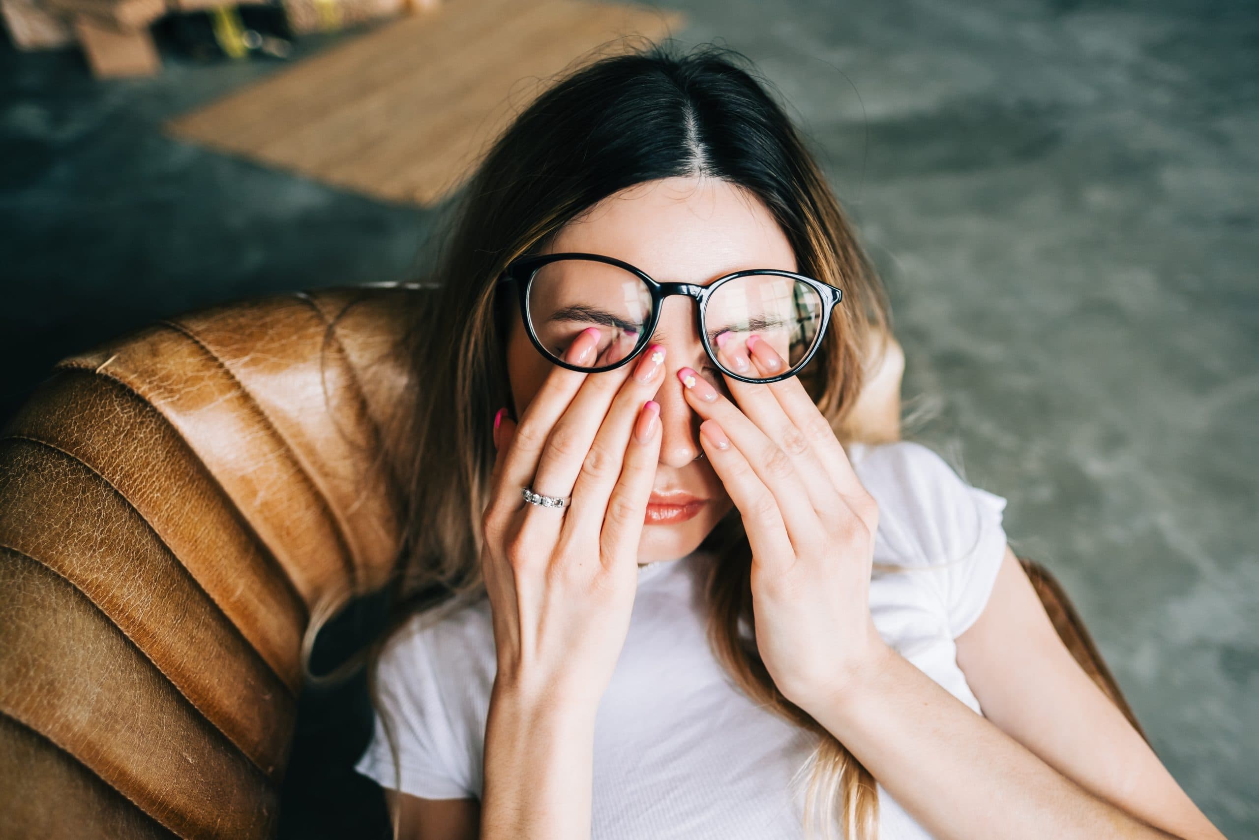 Young woman sitting in armchair rubs her eyes with tiredness, pushing up her glasses.