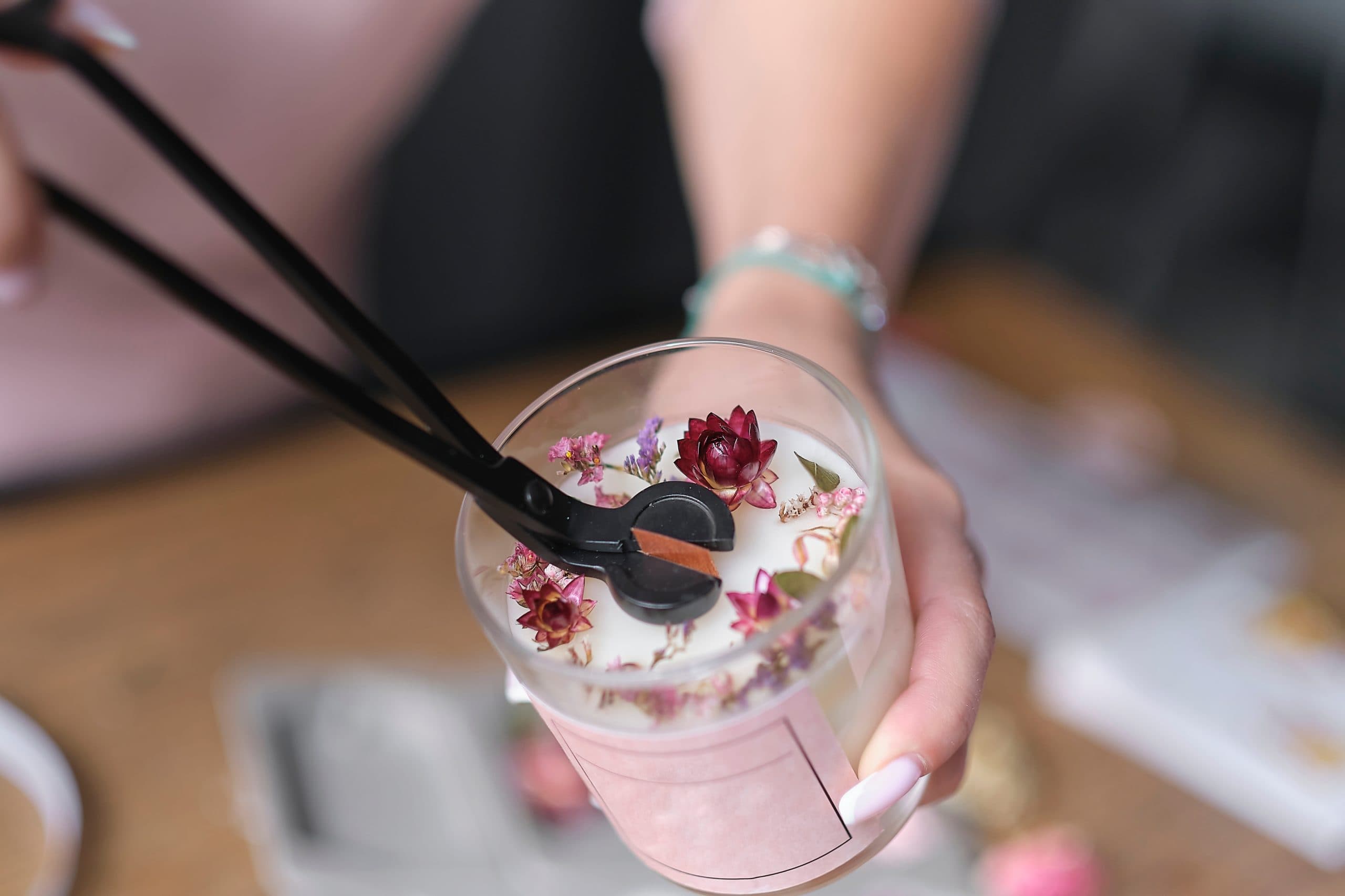 Close up of a newly-made white soy candle with red and pink dried flower decorations. A woman's hand is trimming the wick of the candle with a tool as a final touch.