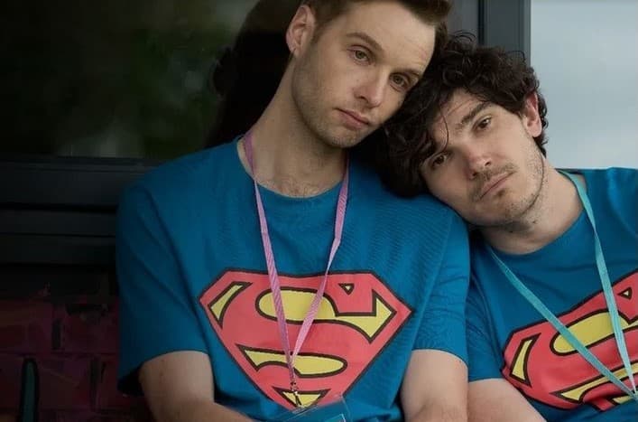 Two young men featured in the TV show "Lost Boys and Fairies", Gabriel and Andy. They are sitting together leaning on each other's shoulders, wearing matching Superman TV shirts and name tags on lanyards round their necks.