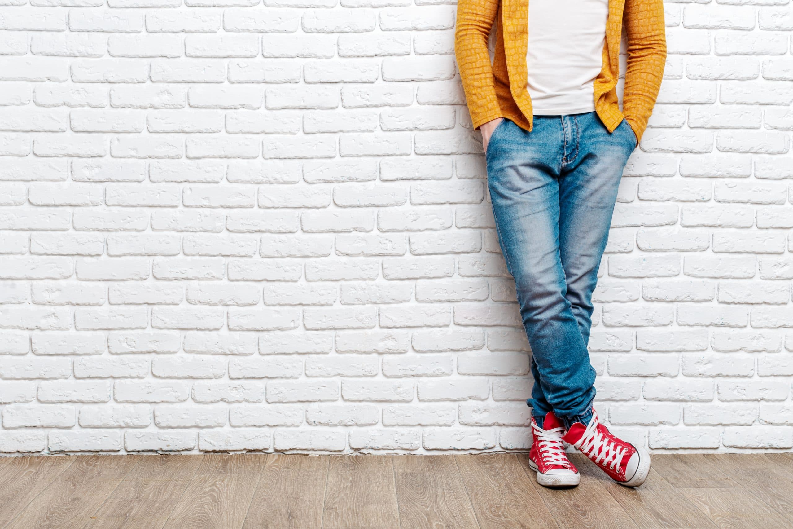 A person seen from the chest down leaning on a white brick wall, wearing blue jeans, red high top sneakers and a dark yellow jacket open over a white top.