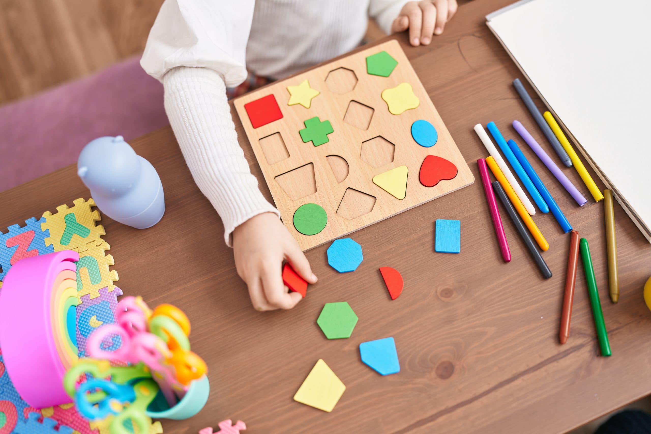 Arms of a child in a light jumper sitting at a wooden table playing with many-coloured shapes and crayons.