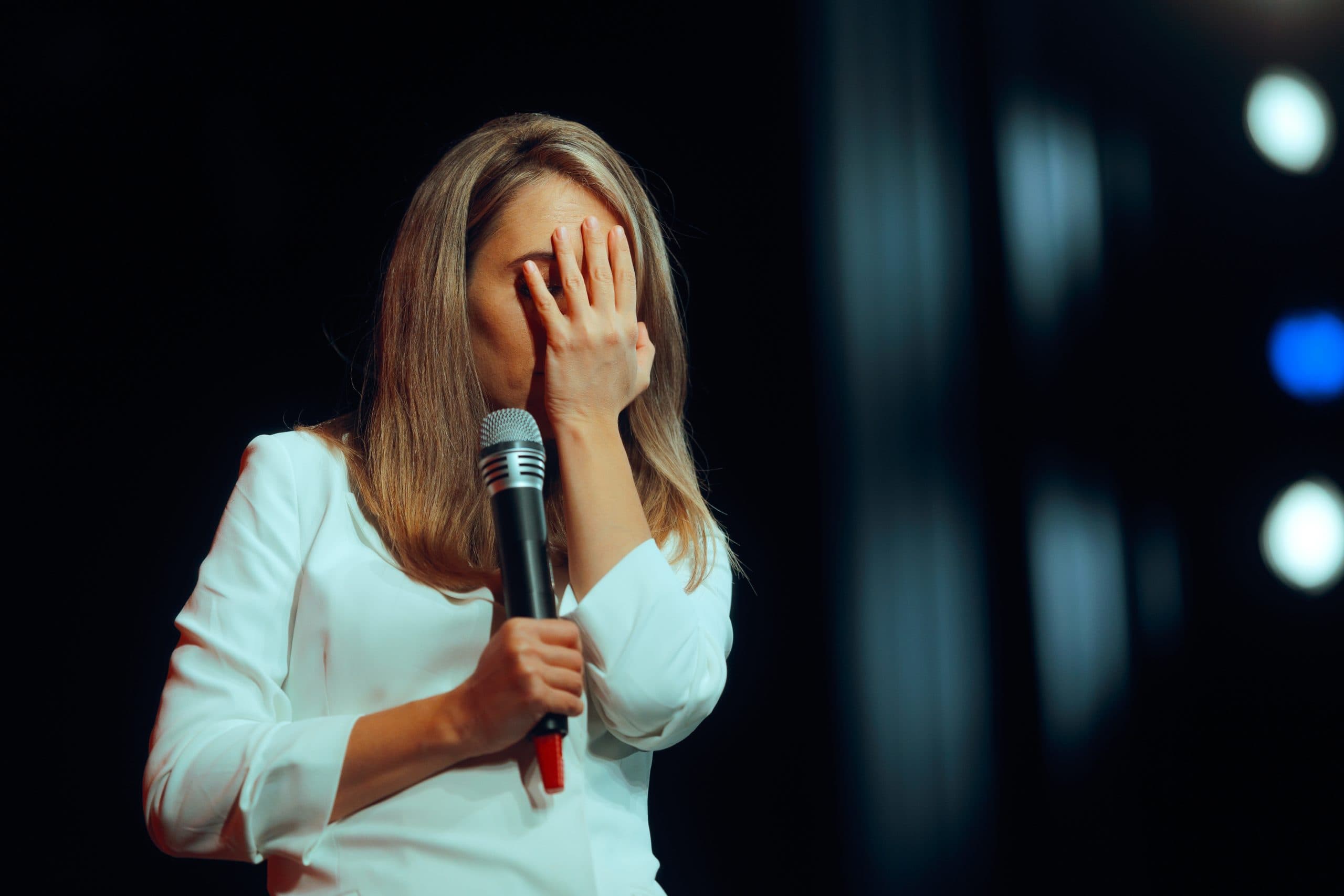 A woman standing nervously with her hand over her face and holding a microphone, surrounded by spotlights.