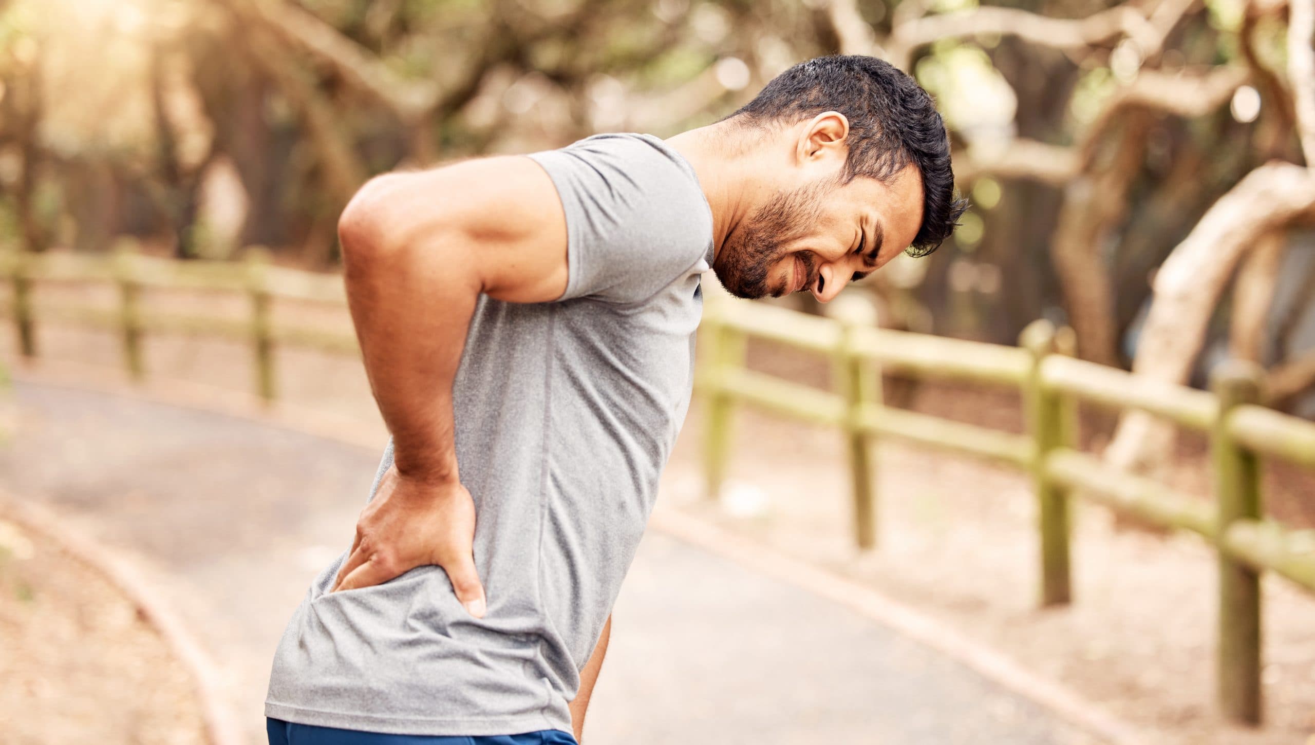 A man in a grey t-shirt and blue shorts is out of doors on a path. He is stopped and bending forward while holding his lower pack. His face shows pain.