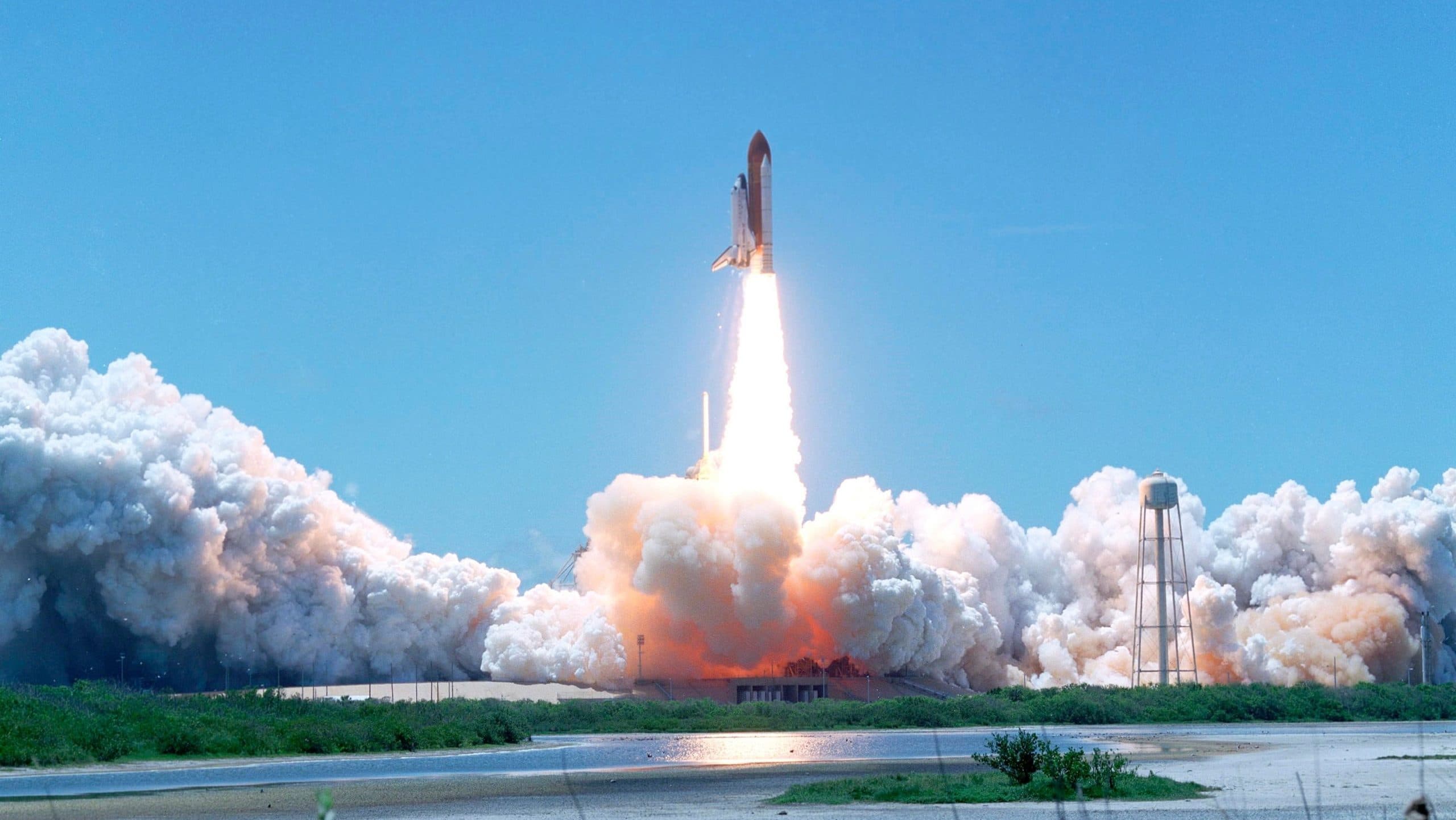 Orange and white smoke and steam billow out across grassland and waters surrounding the rocket launch pads as a space shuttle lifts off. Taken at the Kennedy Space Centre in Florida USA.