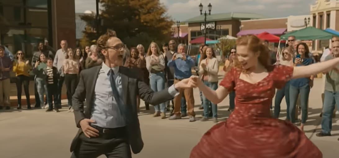 Scene from the movie The Life of Chuck, with the hero Chuck as a young man wearing a suit, and dancing with his friend Janice, wearing a red dress. They are in a public area surrounded by people looking on.