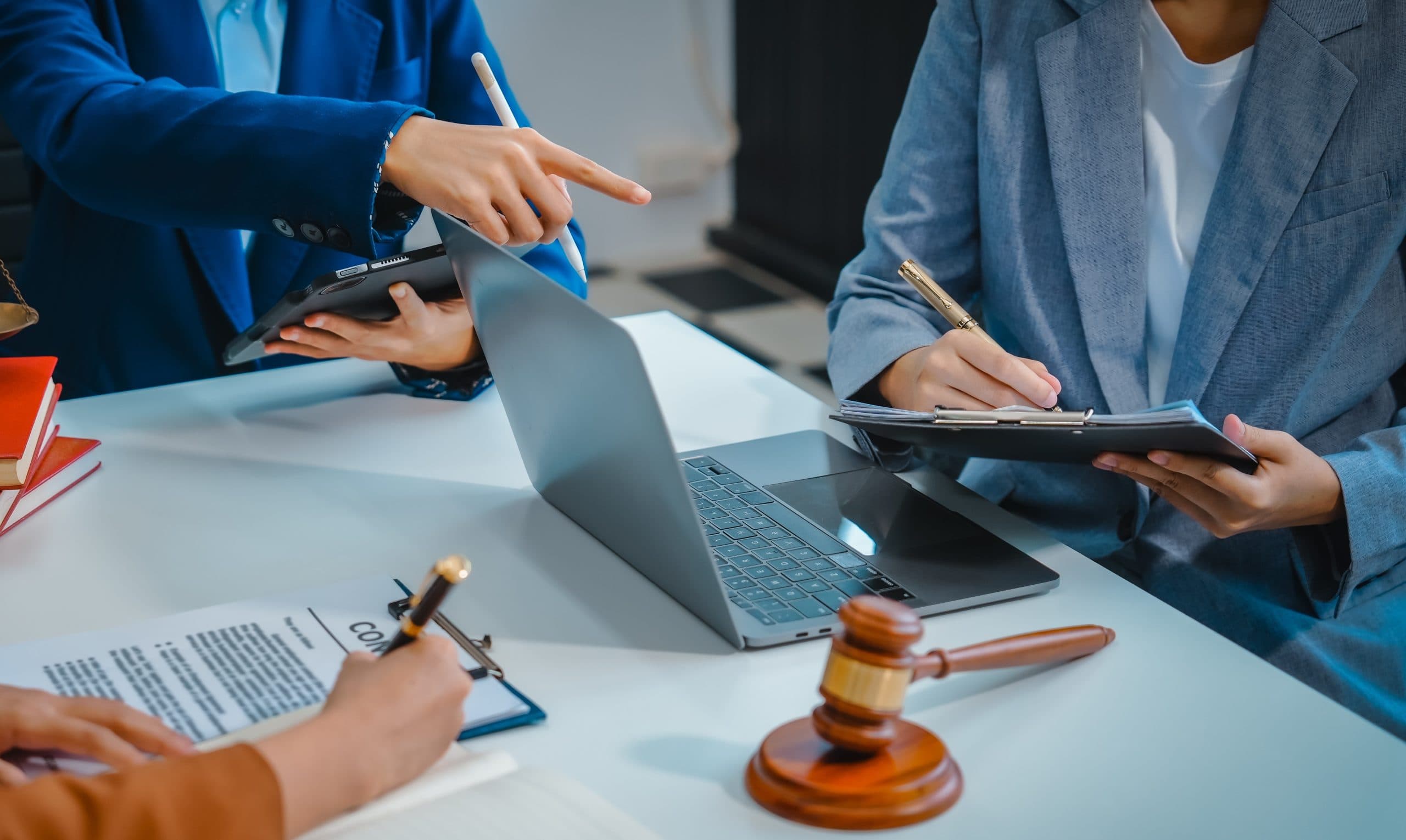 Hands and arms of three people sitting at a table with a contract document, reference books and computers. A wooden legal gavel is on the table in front of them. Two are holding pens. Their gestures suggest they are debating what's in the document and on the computers.