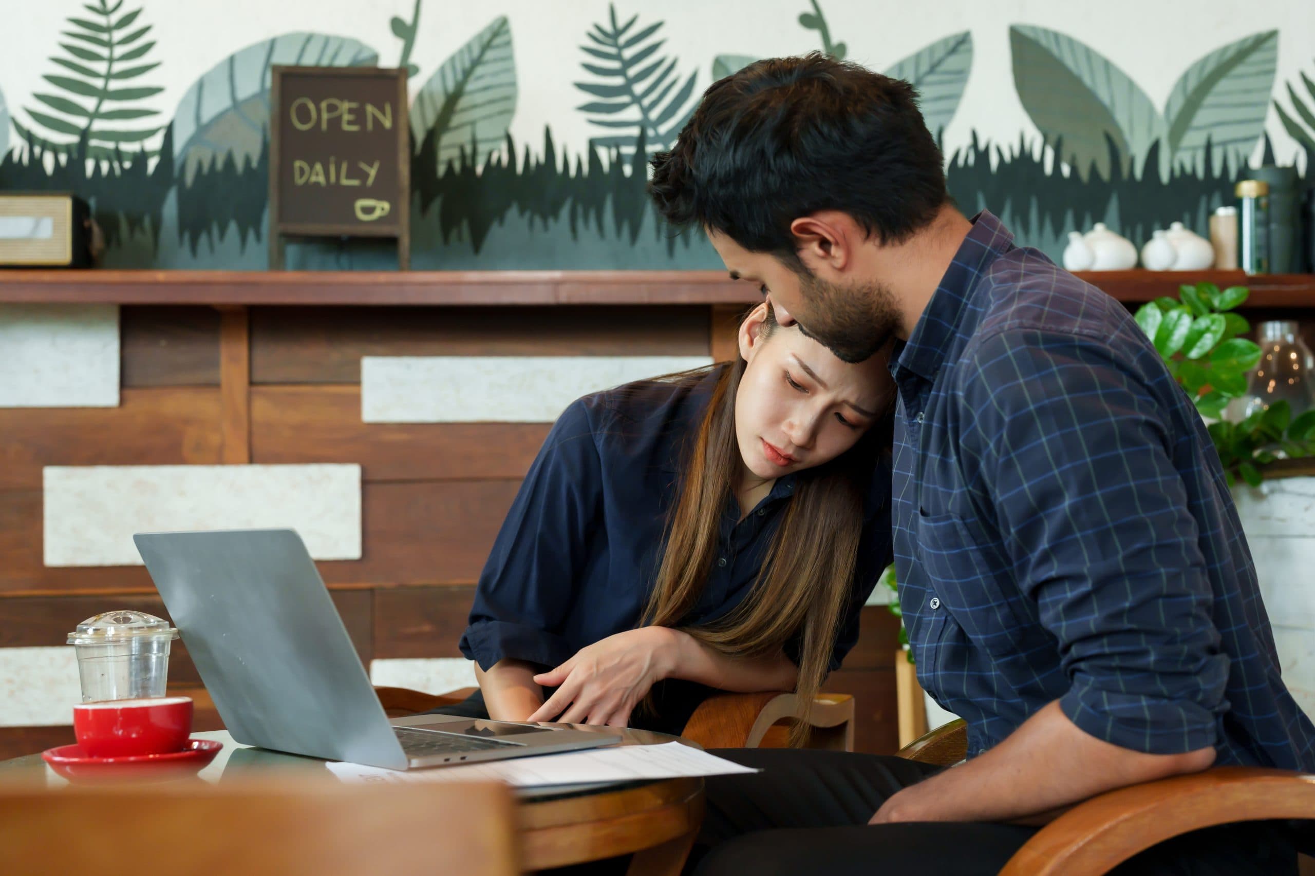 A young man and woman who are owners of a small family business, sitting in their cafe, comforting each other while looking sadly at a laptop and printouts.
