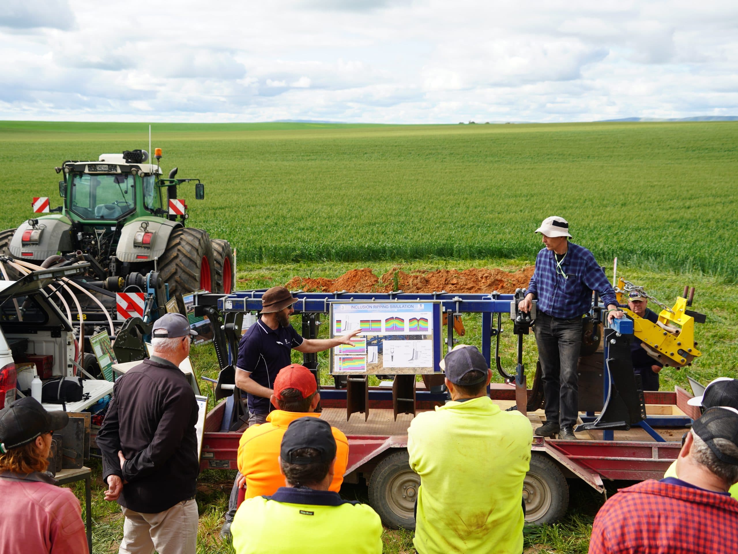 A group of people who are farmers and engineers stand in a crop field and alongside a tractor. There is a white board propped up on equipment demonstrating improved techniques, with two people instructing from it, and around six or seven people looking on.