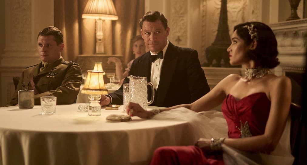 Characters from TV show Rogue Heroes Paddy Mayne (Jack O’Connell), Dudley Clarke (Dominic West), and Eve Mansour (Sofia Boutella), in evening dress, seated at a dining table in a dimly lit restaurant.