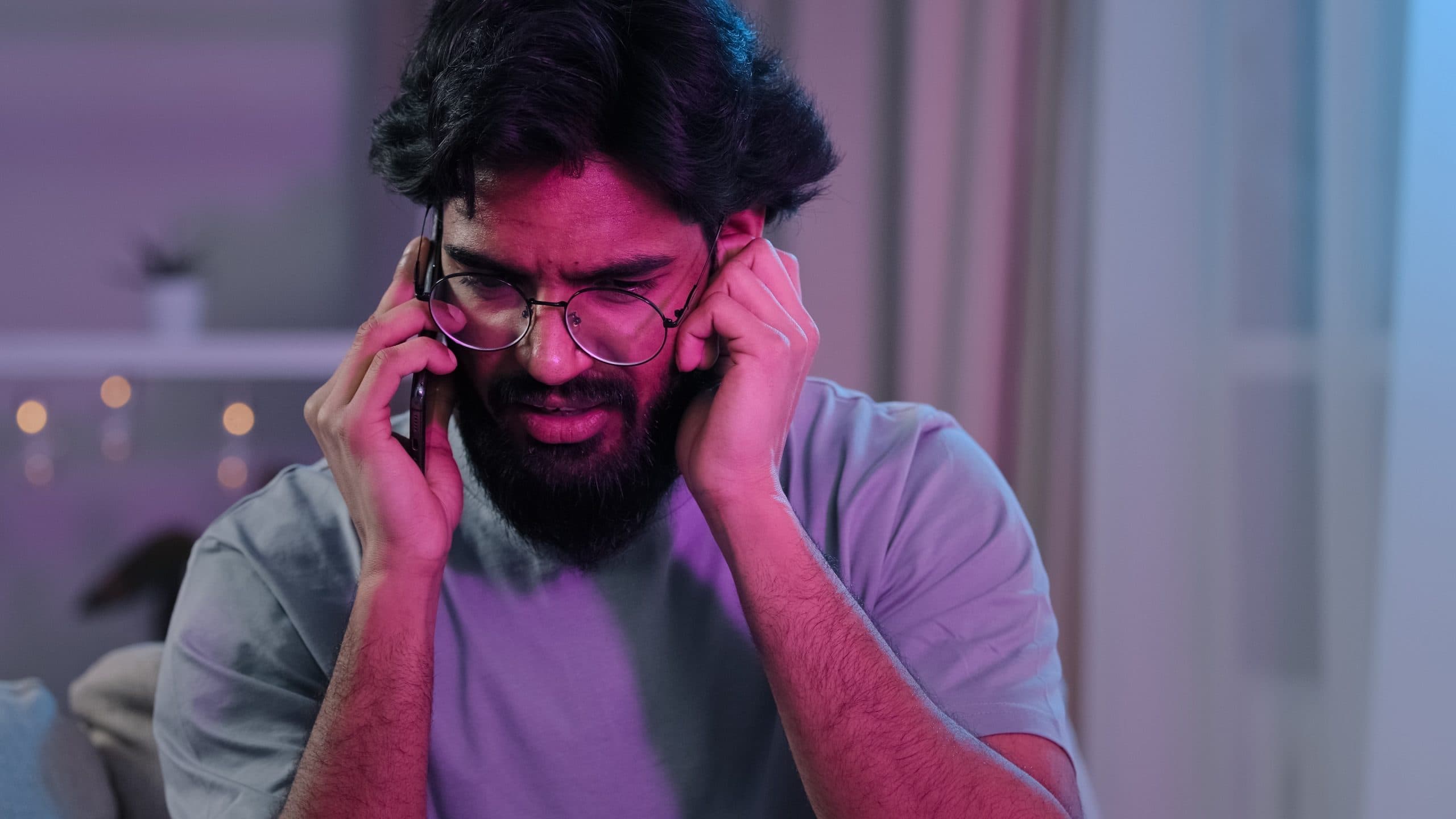 A bearded man with glasses is frowning as he has a hard to hear conversation on his phone in a noisy room.