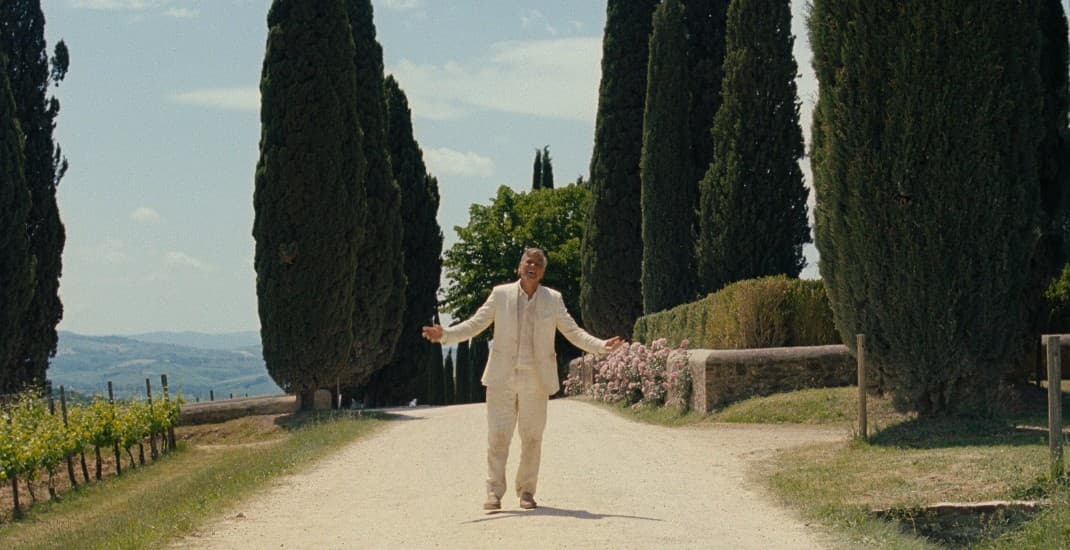Scene from the movie Jay Kelly. The lead played by George Clooney stands with open arms on a gravel drive with pine trees and roses behind. He is wearing a white suit stained with red dust.