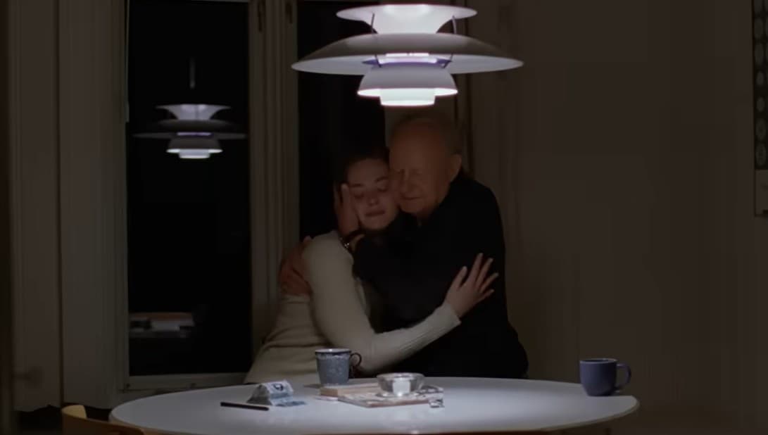 Shows characters from the film Sentimental Value. Daughter Nora and father Gustav embrace, sitting side by side at a round table, in a dim room, with two coffee mugs and some small dishes in front of them on the table.