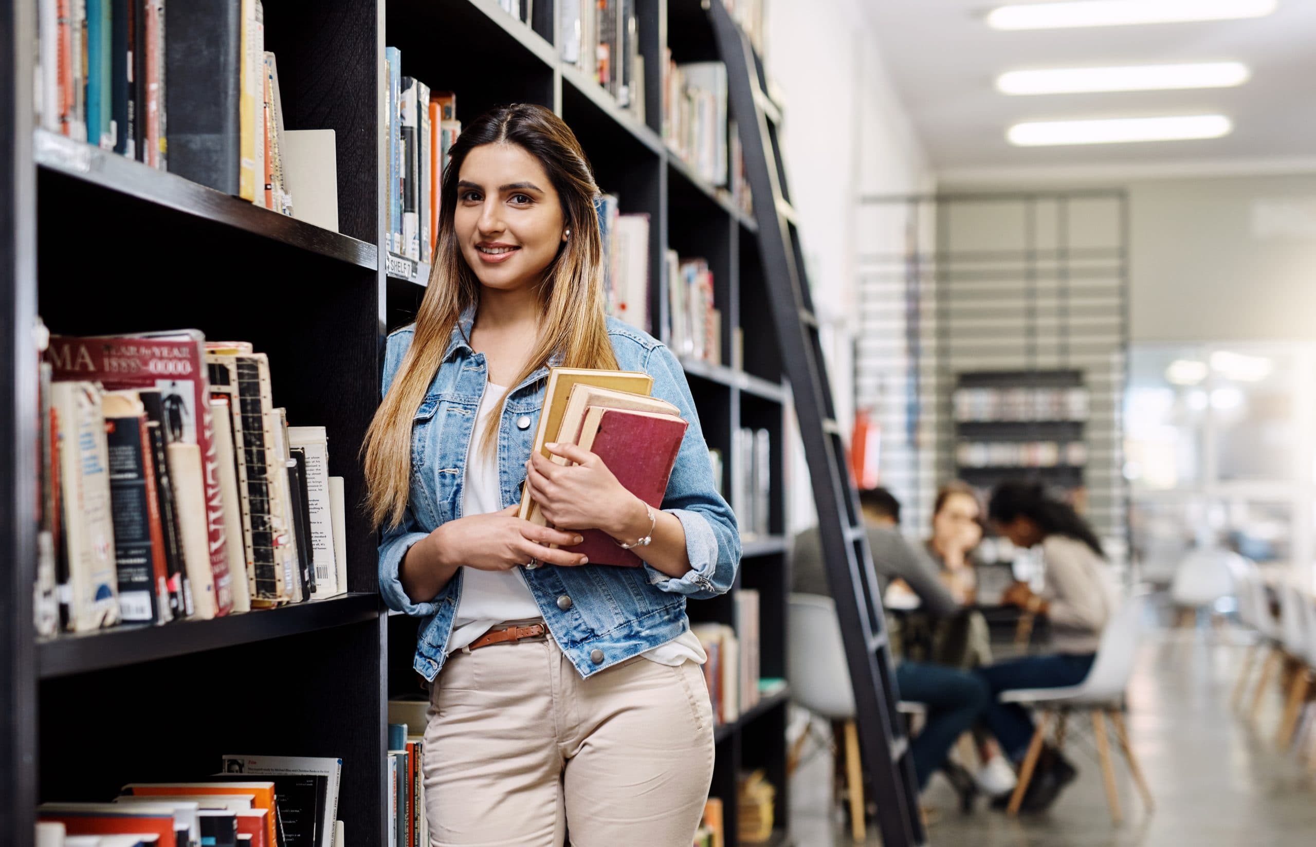 Relaxed young woman in casual clothes leaning on shelves of books in a public library. Behind her in the distance are other people seated around tables, some reading some chatting.